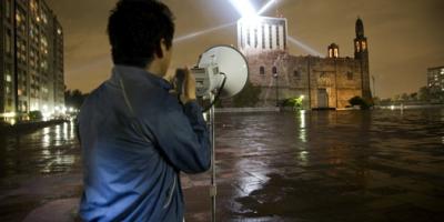 Persona de espaldas que sostiene un megáfono, de fondo el ministerio de Relaciones exteriores de México  y sobre su techo tres haces de luces. 
